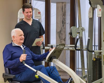 Senior man exercising with staff assistance in facility gym.