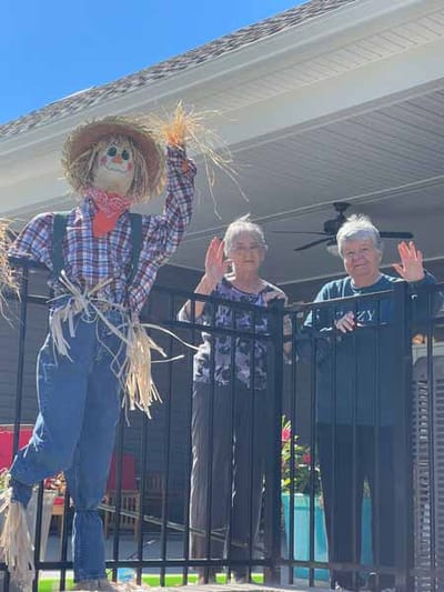 Two residents waving with a scarecrow outside.