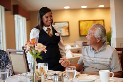 A staff member serving a meal to a smiling resident in a dining area