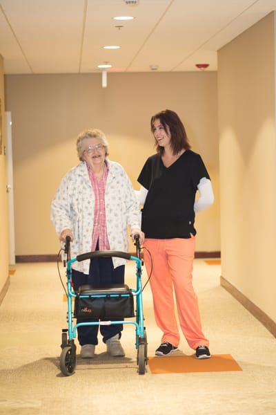 A senior resident with a walker accompanied by staff in a hallway
