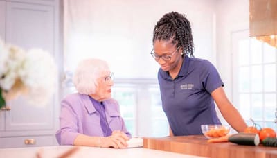 A caregiver and a senior woman engage in conversation in a bright kitchen setting.