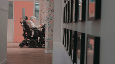 A resident in a wheelchair in a hallway with framed photos