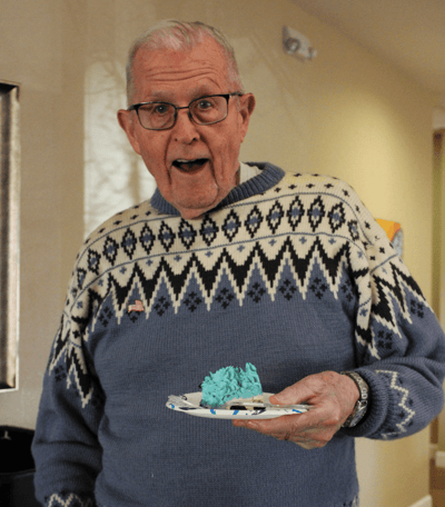 An elderly man in a sweater smiling with cake.