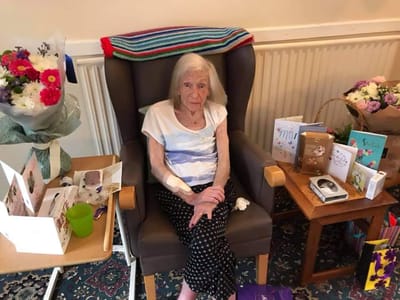 Elderly woman sits in chair among decorations.