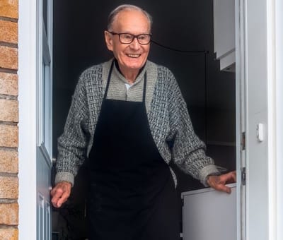Elderly man smiling at the door wearing an apron.