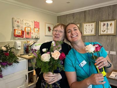Staff member and resident smiling with flowers in a common area