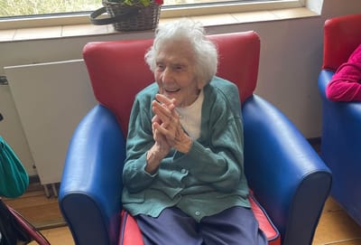 Elderly woman smiling while seated in a blue chair.