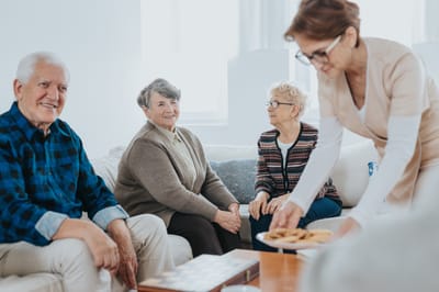 Seniors chatting in a cozy common area with a caregiver serving snacks.