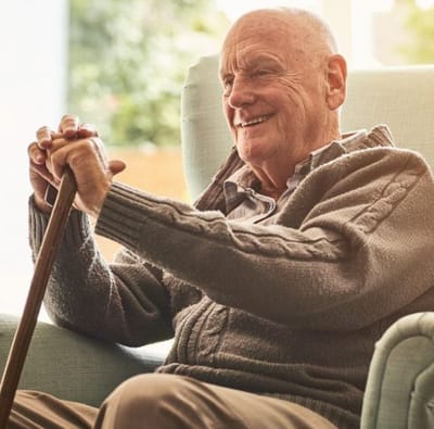 An elderly man sitting in a chair, smiling and holding a cane.