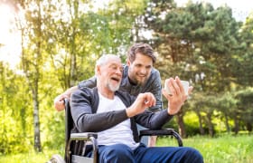 Smiling elderly man in a wheelchair with a young man using a smartphone outdoors