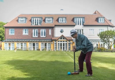 An elderly man playing croquet on the lawn in front of the building.