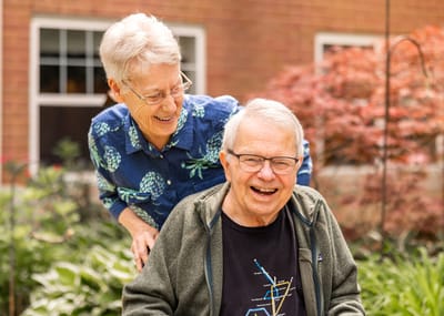 A woman pushing a man in a wheelchair, both smiling outdoors