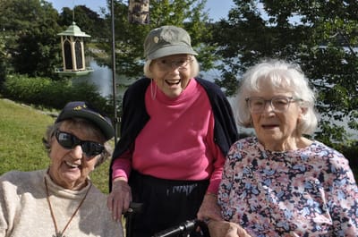 Three smiling women by a lake with greenery.