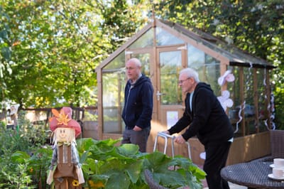 Two residents walking in a garden area near a greenhouse
