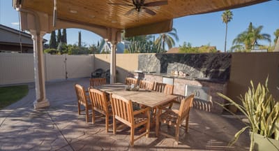 Wooden dining table and chairs under a shaded patio with an outdoor kitchen.