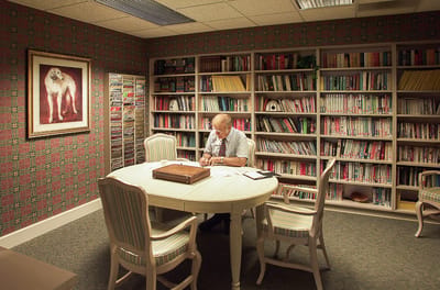 An elderly man reading at a table in a library-like room