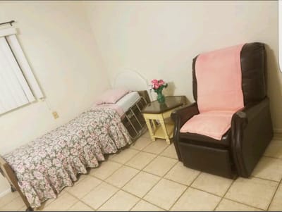 A small bedroom featuring a floral bedspread, a reclining chair, and a small table with flowers.