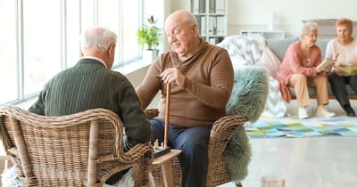 Two seniors playing a game indoors, with others reading.