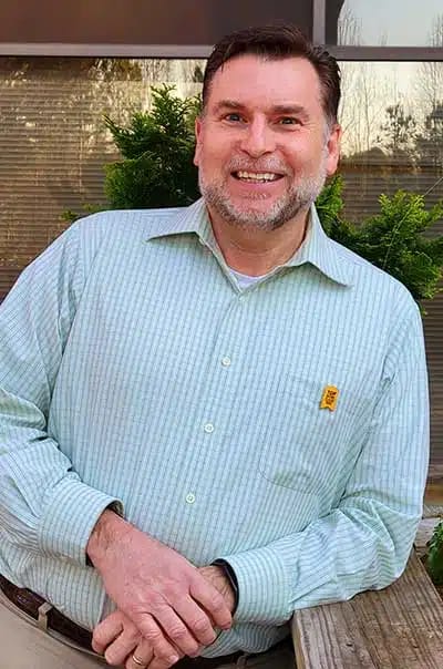 Man smiling beside greenery in an outdoor setting.