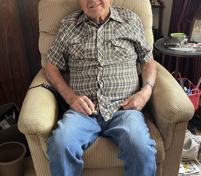 Smiling elderly man in plaid shirt sitting in an armchair.