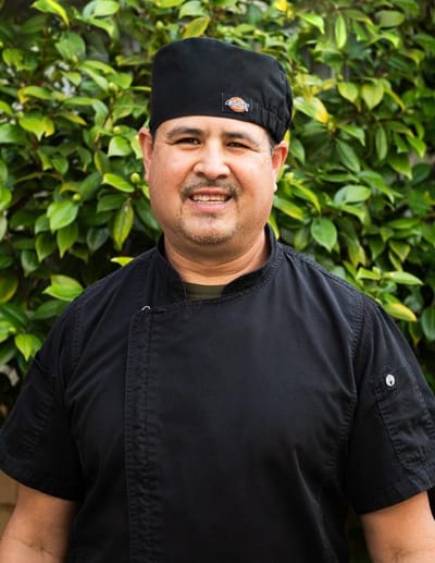 A chef smiling in front of a green bush