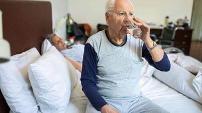 An elderly man drinking water while sitting on a bed