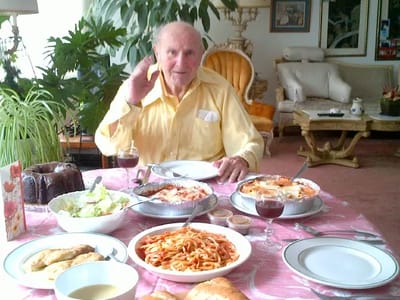 An elderly man waving at a dining table with food