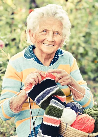 Elderly woman knitting outdoors in a garden.
