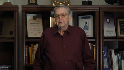 An elderly man stands in front of bookshelves with awards.