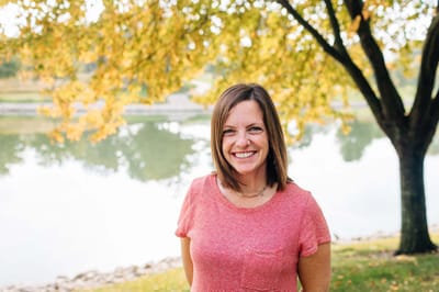A smiling woman standing by a pond with fall foliage