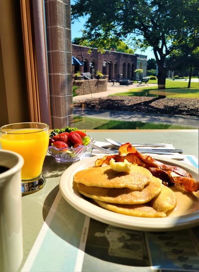 A breakfast plate with pancakes, bacon, and juice by the window