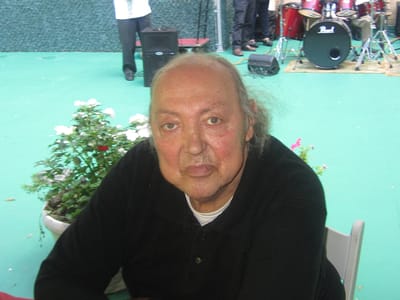An elderly man seated outdoors, surrounded by flowers
