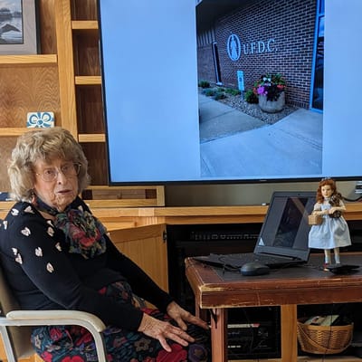 Elderly woman seated in a common area with a screen showing the building.