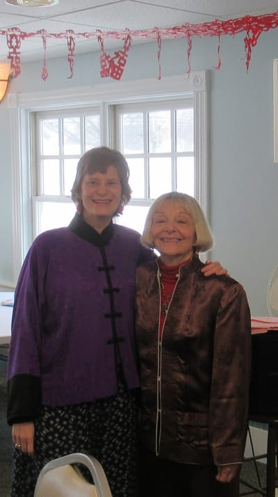 Two women in winter clothing smiling together indoors.