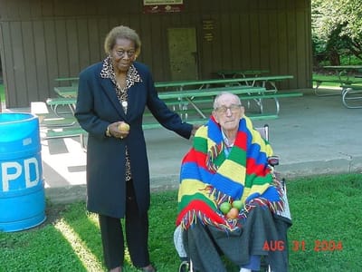 Elderly man in wheelchair with colorful blanket and woman outdoors.