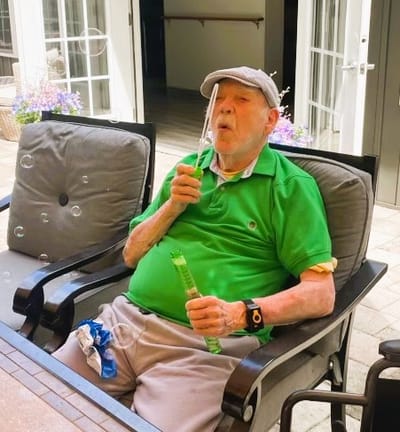 Elderly man blowing bubbles outdoors in a chair.