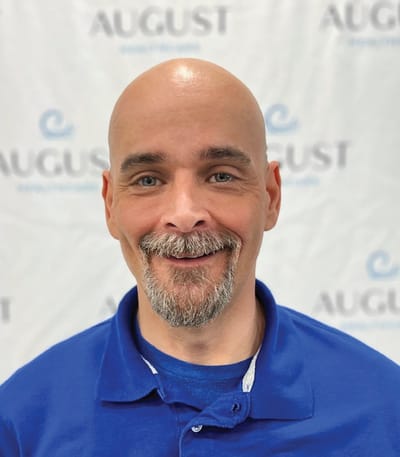 Staff member smiling in front of August Healthcare backdrop.