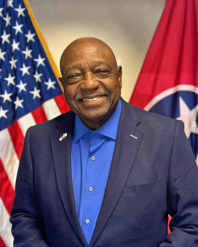 Smiling man in a suit with flags in the background.