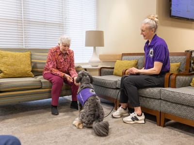A resident interacts with a therapy dog and staff member indoors