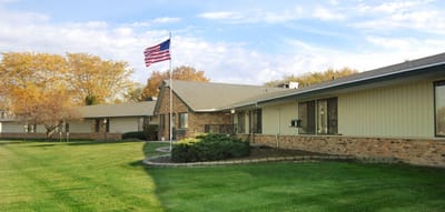 Alden Meadow Park Health Care Center, Inc, Nursing Home in Clinton, WI — Senior living facility with a flag and landscaped grounds