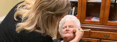 A caregiver applying makeup to a resident in a cozy room