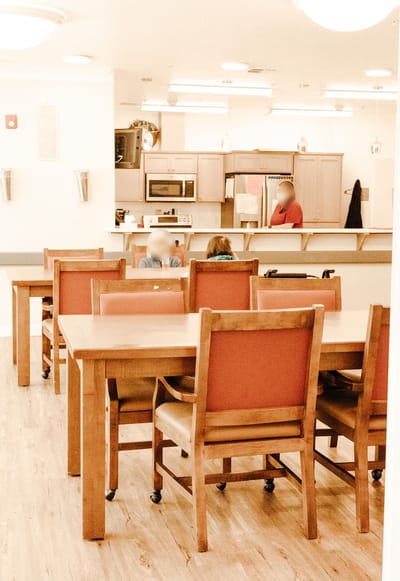 Bright dining area with residents and kitchen in the background
