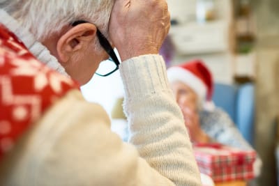 Close-up of a senior man holding his glasses, with a woman in the background