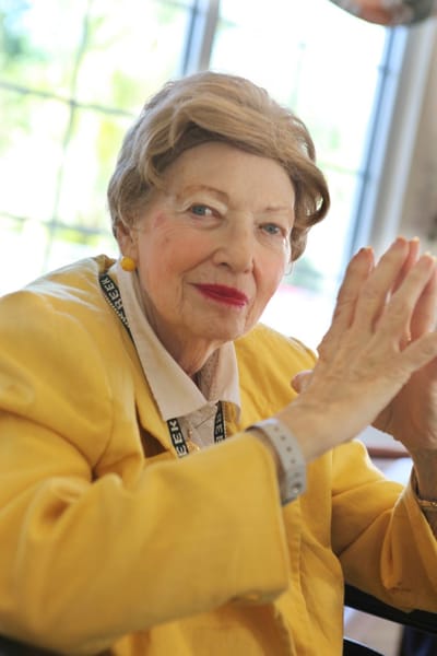 A senior woman in a yellow jacket with clasped hands