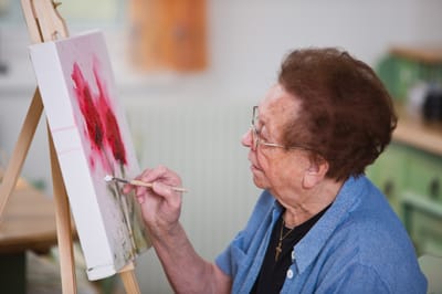 An elderly woman painting on a canvas in a bright room