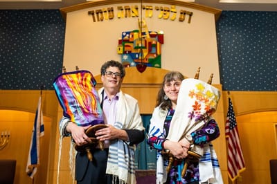 Two individuals holding decorated Torah scrolls in a facility interior