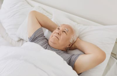Elderly man resting in bed with hands behind head.