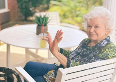 Elderly woman smiling and waving at a table.