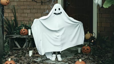 Child dressed as a ghost near pumpkins in an outdoor setting.