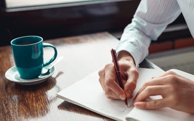 Person writing in a notebook with a coffee cup.